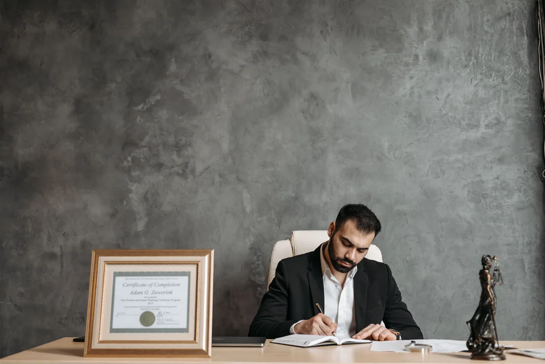 Lawyer writing at a desk beside a framed certificate and a lady justice statue